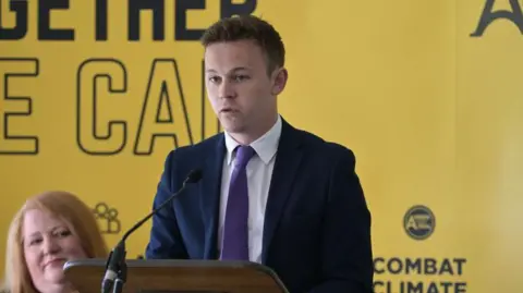 Getty Images A man is speaking into a microphone. He is wearing a dark suit, white shirt and purple tie. He is standing in front of yellow wall which is branded with Alliance Party slogans.