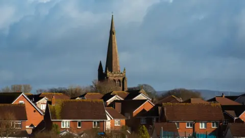 A church spire rises above several houses in Millom. Hills can be seen in the distance.