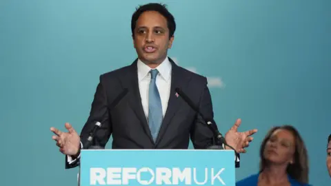 PA Media Former Reform UK Chairman Zia Yusuf is pictured wearing a black blazer, white shirt and blue tie. He is stood in front of a lectern with a Reform UK sign