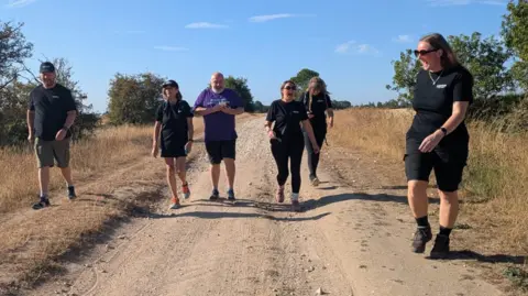 Six people walk towards the camera along a dusty footpath with dry-looking grass on both sides and a smattering of green bushes. The sky is blue with wispy clouds. On the right, nearest the camera, a woman in a black T-shirt, shorts and sunglasses is laughing.