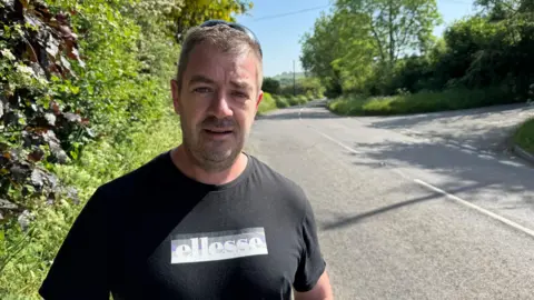 A man with short hair and sunglasses pushed up on his head stands at the edge of a country road. He is wearing a black t-shirt with an Ellesse logo across the chest. Green hedgerows and trees line the road behind him under a bright blue sky.