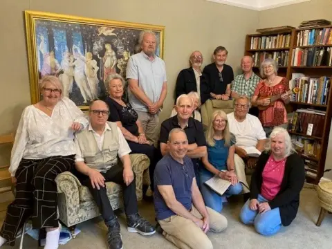 BBC / Sally Fairfax Fourteen men and women, all over 50 years old, sitting in a group in a book-lined living room, smiling