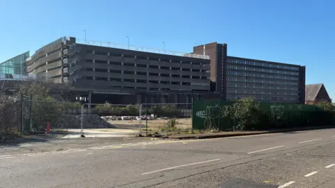 Peter Cooper/BBC A multi-storey car park positioned next to a vacant office building site - Belgrave House - which is of a similar size in height. The buildings are viewed from Lady's Lane in Northampton on a clear day and viewed across the derelict site of the former Greyfriars bus station