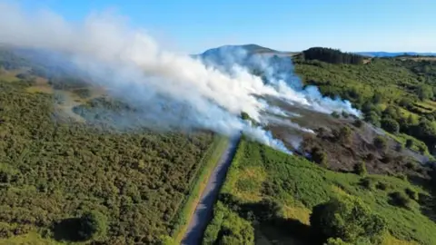 An aerial view of hills, with various pockets of smoldering bush sending white smoke high into the blue sky