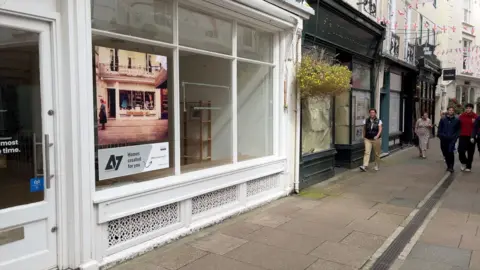 BBC Empty shop fronts in Mill Street, St Peter Port, Guernsey. People can be seen walking on the right side of the image while a white empty shop is on the left with a boarded up green shop front next to it