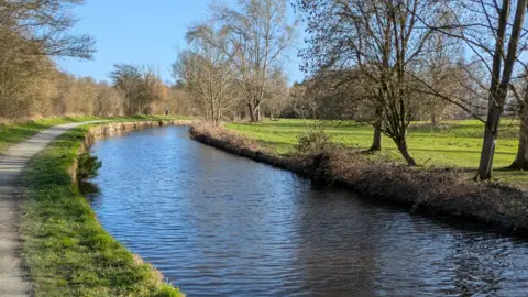 A towpath is on the far left of the photo, by grass, which is next to the canal. A field is to the right of the canal and trees are to the left and right of the canal.