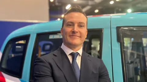 A man in a shirt and tie standing in front of a taxi painted in Reform UK colours. He is smiling at the camera.
