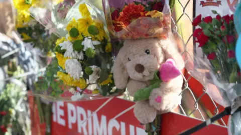 A bouquet of flowers and a soft toy dog are attached to a fence at the tribute site above a red and white sign.