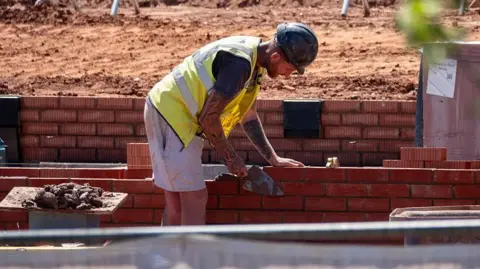 Getty Images A construction worker in a yellow hi-vis vest and grey shorts is laying red bricks on a partially built brick wall, using a trowel to apply mortar.
