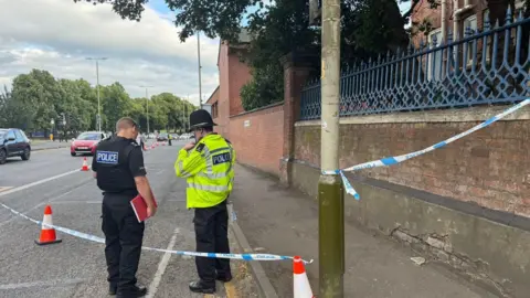 Leicester Media Online Two police officers standing by a tape cordon on the edge of a busy road.