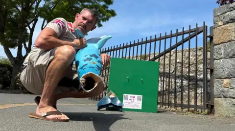 Steve Byrne is crouching down outside the school next to the plinth. He is holding the dolphin model to show the its tail has been ripped off. On the plinth is the remains of the tail. 