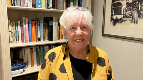 Elspheth Davis stands in front of a book shelf in her home. She has short grey hair with a pair of glasses on top of her head. She is wearing a yellow shirt with black circles and a black top underneath.