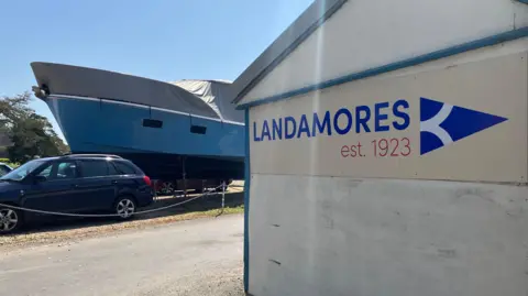 Owen Sennitt/BBC A sign saying Landamores is attached to a white building with a large blue boat in the background. 