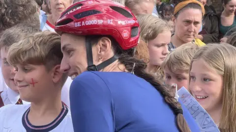 BBC Scott stands in cycling gear and helmet with children and adults who have come to see her on her challenge. She is posing for photos with children who are smiling. She has long brown hair in a plait, emerging from under a red cycle helmet, and is wearing a blue cycle top. 