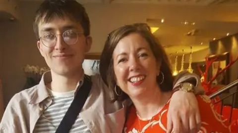 Anne-Marie Hoare Head and shoulder image of Anne-Marie Hoare stood next to her son Marley, who has his arm around her. Anne-Marie has shoulder-length brunette hair, is wearing hooped earrings and a red top. Marley has short brown hair, round glasses and is wearing a beige jacket over a white and grey stripped top.