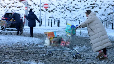 A woman pushes a trolley full of her supermarket shopping through far-stretching, thick brown slush in a car park. She wears an ankle-length grey winter coat and her trolley is full to the brim with bagged and unbagged shopping items including a large bottle of milk. In the background, a couple wearing dark clothing push their own shopping trolley towards their car, and beyond them are snow-covered trees. 