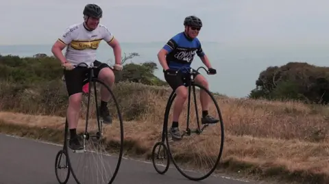 Two men in cycling Lycra and helmets pedalling penny farthings on a road near a cliff edge.
