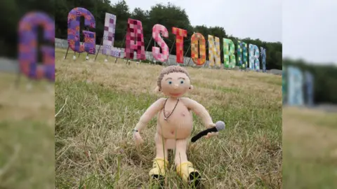 Jason Bryant A knitted doll resembling Chris Martin standing on the grass in front of the colourful Glastonbury sign. He is naked and holding a microphone and wearing yellow boots