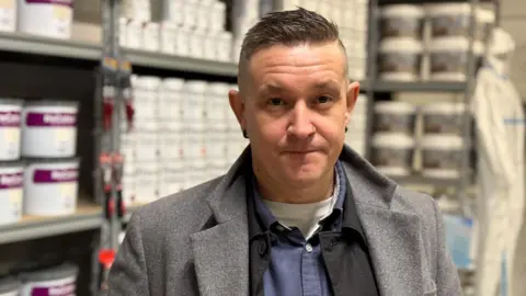 BBC A man with short, dark, spiked hair looks at the camera. He has a nose ring and dark earrings. He is wearing a grey coat, a blue shirt and a white vest. Many tins of paint are in the background on shelving.