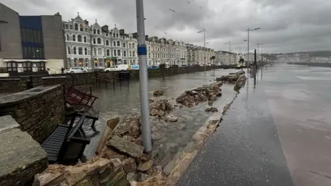 MANX RADIO The Sunken Gardens on Loch Promenade, which has a section of stone wall that has been knocked down by waves. The garden space is filled with water and two wooden benches have been knocked over. The promenade walkway stretches out into the distance on the right, while the tall Victorian Buildings on the promenade can be seen on the left.