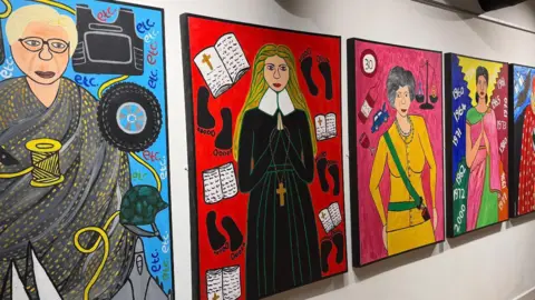 The photo shows four colourful paintings hung up on the wall. They all depict women, and have been painted in bright colours. 