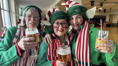 Three women dressed in green, white and red elf outfits holding a drink. 