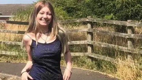 Family Handout Morgan Dorsett wearing a dark blue dress with silver sequins forming a pattern on the front. She is smiling at the camera, and has long blonde hair with darker roots. She is wearing black eyeliner, a choker necklace and bracelets on each wrist. Behind her there is a curving pavement and a wooden fence, overlooking a patch of dry grass and a large green bush on a sunny day.
