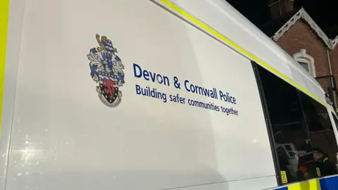 BBC A white police van with the police emblem on the left, the Devon & Cornwall police title to the right and the saying 'Building safer communities together' underneath. A house can be seen in the top right corner.