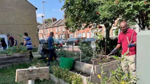 Edible High Town A group of adults garden raised beds at the end of a terraced street. It is summer and they are holding tools and seedlings.