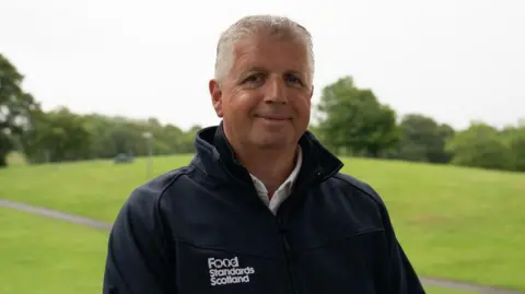 A man with white hair wearing a black jacket with "Food Standards Scotland" emblazoned on it smiles at the camera. He is standing in an open green space like a park