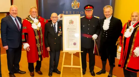 Dudley Council Six men standing in a room, two with red ceremonial robes, three in suits and one in military uniform, with a large certificate on a stand in the middle