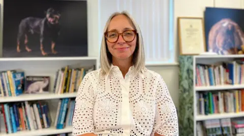 Pam Aubert stands in front of a number of book cases. She is wearing a white top, has shoulder-length blonde hair and is wearing glasses.