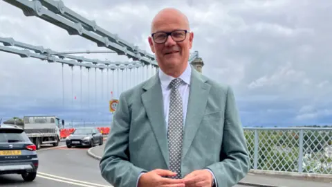 Dale Spridgeon A middle-aged bald man in a grey suit stood outside a large bridge crossing with cars travelling in both directions.