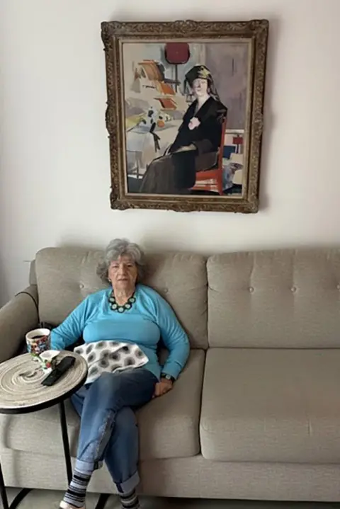 PA Media Helene Plotkin wearing a blue jumper and jeans sits on a grey sofa with the painting hanging on the wall behind her.
