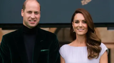 Getty Images Prince William and the Duchess of Cambridge standing on the green carpet at the Earthshot awards in 2021. William is wearing a dark green velvet jacket and a navy blue roll-neck sweater. Kate has a simple white dress on. Both of them are smiling at the camera.