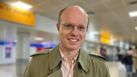 Adam Boggon smiles for the camera - he is wearing a light green jacket, has glasses and is balding.