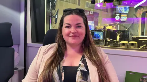 Sue Dougan/BBC Heather Williams stares at the camera, she has long dark hair, wears a beige jacket and a black top. She is sitting in front of a radio studio. 
