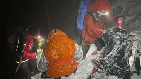 Llanberis Mountain Rescue Team rescuers with ruck sacks in heavy driving snow with a casualty sitting in snow in a plastic rescue bag