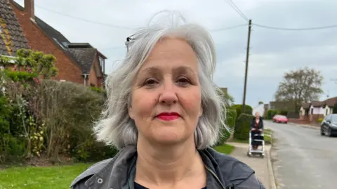 Simon Dedman/BBC A smiling woman looks straight at the camera while standing on a pavement. She is wearing a grey coat.