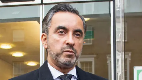 PA Media Headshot of solicitor Aamer Anwar. He has a beard, is wearing a suit and a dotty tie. He is standing in front of glass panelled doors.  