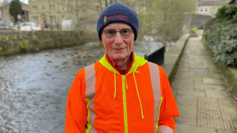Mark Ansell/BBC Andrew is wearing a high visibility orange jacket and blue hat and glasses. He is standing on the towpath next to the River Calder.