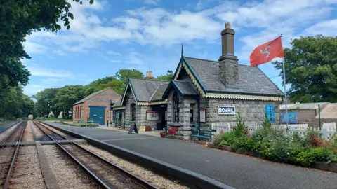 GRANT TAYLOR The Castletown Station made of grey stone, comprises two buildings, with railway lines in front of it.