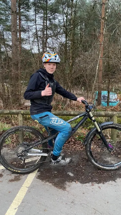 Family photo Side-on photo of 13-year-old Ioan in a forestry car park, wearing a helmet and cycling clothing, sitting on his bike and giving a thumbs up.