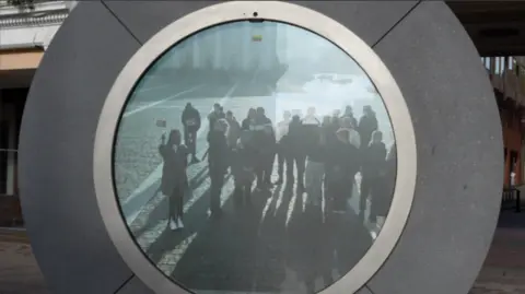 John Fairhall/BBC A circle portal stood in the middle of the image. It is made out of silver material and has a screen in the middle showing people standing in it. 