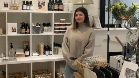 
Ellie Schreibke, Owner of Maison Lifestyle. She is young, with brown hair and a small purse lipped-smile. She is wearing a high neck brown nit jumper and blue jeans with her arms cross ahead of her. She is stood in her shop, surrounded by homeware that has a beige tone. 
