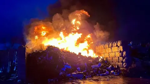 A large fire on a pile of metal that is contained in a constructed wall. The scene is dark.