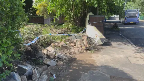 A front garden is taped off. There is rubble and ripped up hedges from a recent crash. A bus can be seen in the distance