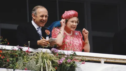 Getty Images Queen Elizabeth II with looking excited with her arms up at the derby races, next to her there's Sir William Heseltine holding a notepad in his hands.