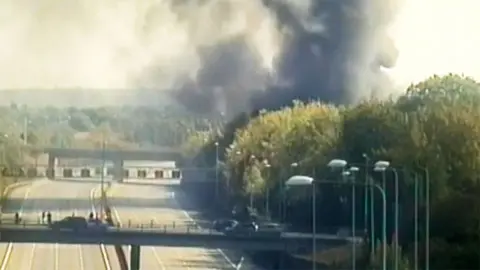 A large plume of smoke by the M25 motorway, with cars on the road across a bridge. Trees are to the left and all around the road.