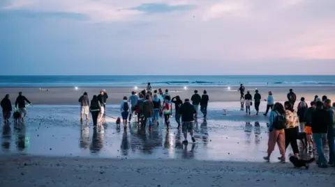 A large group of people walk along a beach towards the sea at sunrise or sunset. Their backs are to the camera.